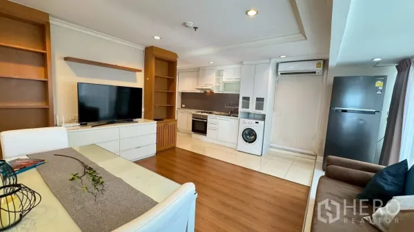 Condominium for rent or sale in Watthana, Bangkok - View of living room showing TV, fitted kitchen with oven and washer, fridge and wide windows.