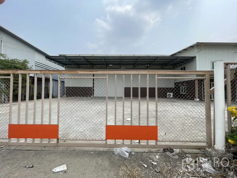 Warehouse for rent in Lat Lum Kaeo, Pathum Thani - Sliding chain-link gate opening to a large concrete yard in front of a metal-clad warehouse.