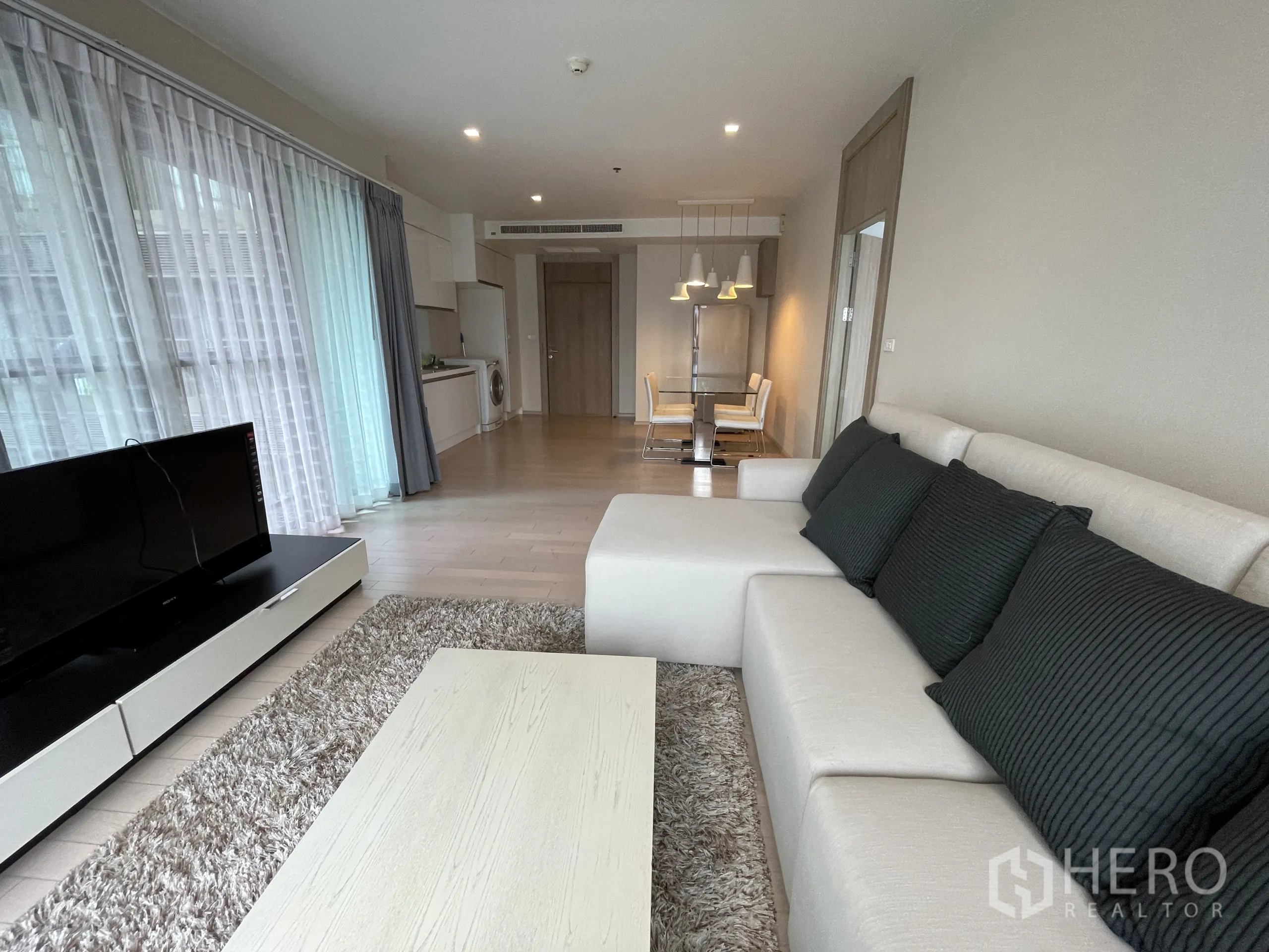 公寓 出租在Watthana, Bangkok - 客厅面向餐厨区，长电视柜、L 型沙发与整面高窗。