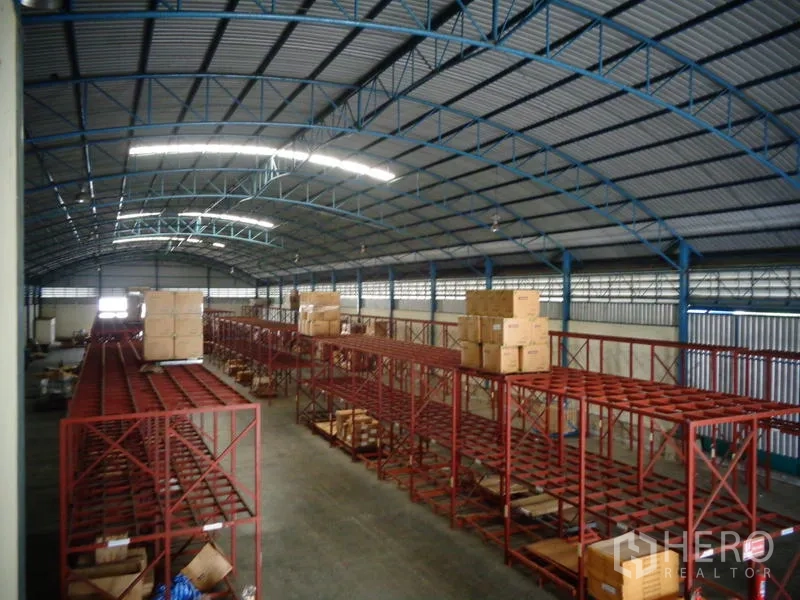 Warehouse for rent or sale in Lat Lum Kaeo, Pathum Thani - High-ceiling warehouse showing blue roof structure and red racking filled with cartons.