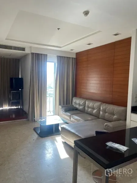 Condominium for rent in Khlong Luang, Pathum Thani - Bright living room with gray leather sofa, wood accent wall, and a balcony door with curtains at Nusasiri Grand.
