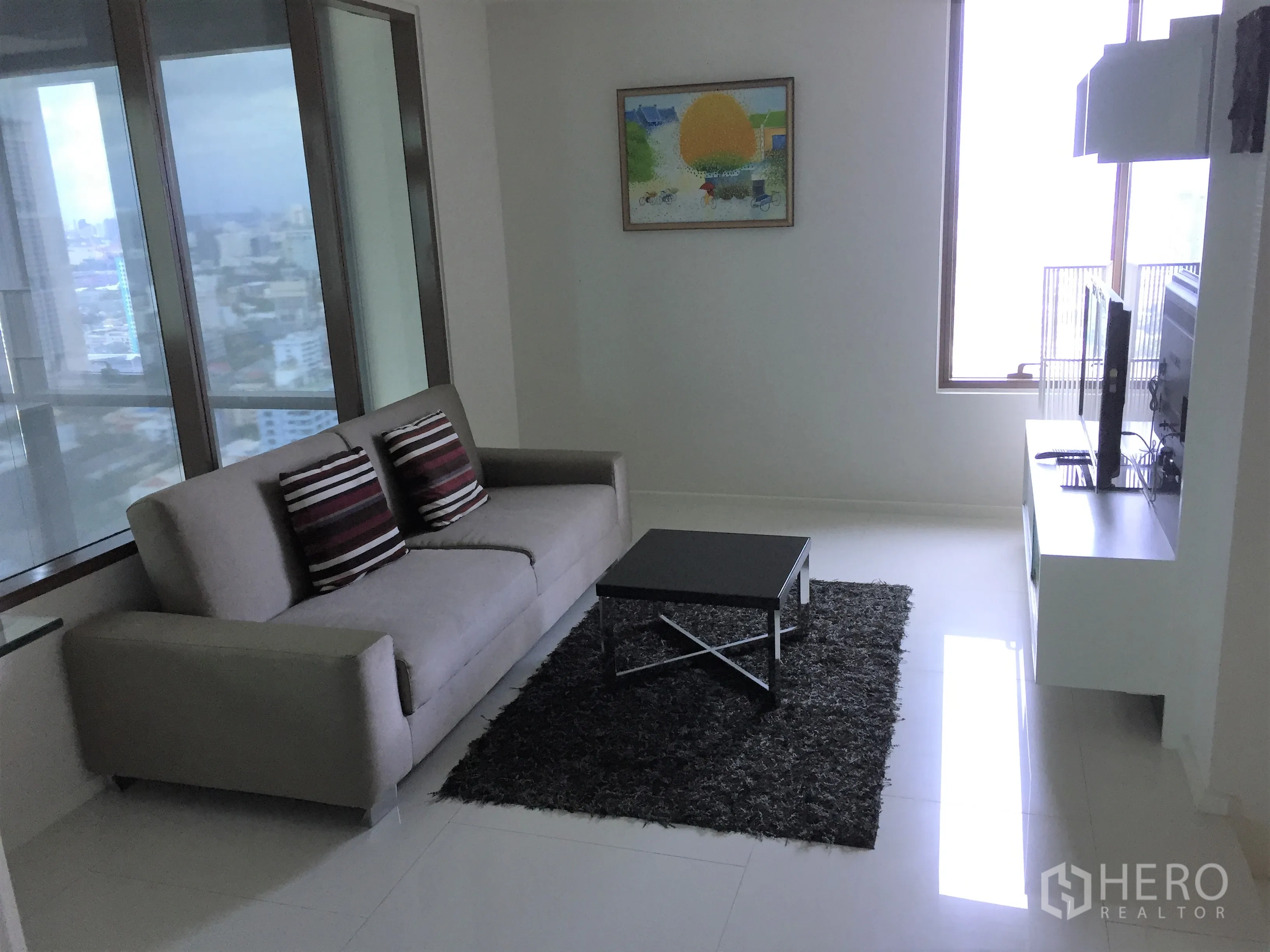Condominium for rent or sale in Khlong Toei, Bangkok - Bright living room with gray sofa, TV console and floor-to-ceiling windows offering city views.