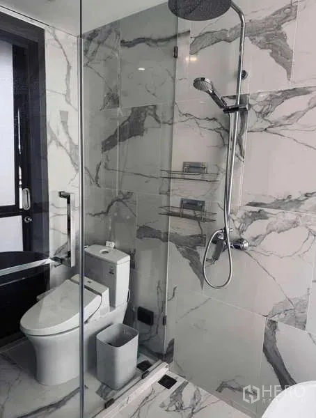 Condominium for rent in Khlong Toei, Bangkok - Glass-enclosed shower and toilet on marble walls in modern bathroom.