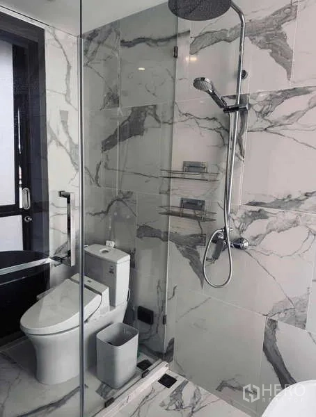 Condominium for rent in Khlong Toei, Bangkok - Glass-enclosed shower and toilet on marble walls in modern bathroom.