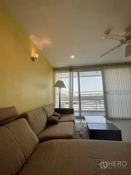 Condominium for sale in Watthana, Bangkok - Living area with L-shaped sofa, floor lamp and sliding glass doors to a balcony with city view.