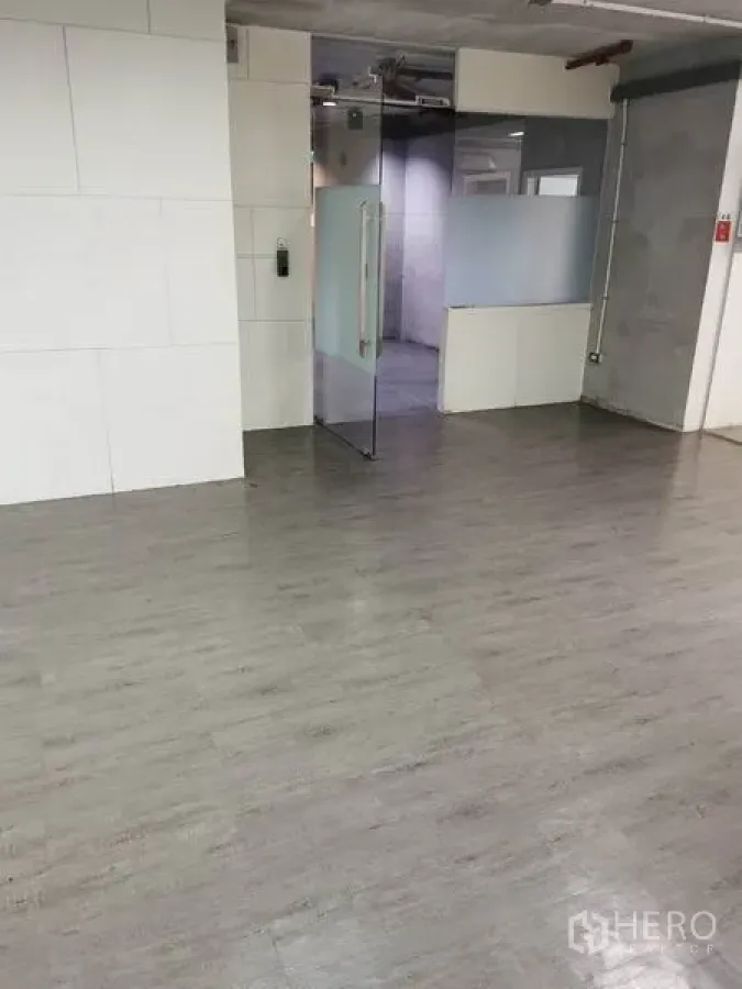 Office Space for rent in Khlong Toei, Bangkok - Open-plan office interior with grey vinyl flooring, white wall panels, glass entry door and frosted partition, plus exposed ceiling piping.