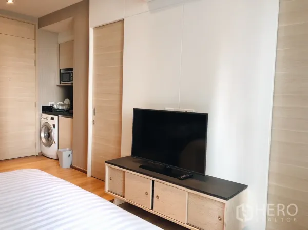 Condominium for rent in Khlong Toei, Bangkok - Bright studio with TV console and sliding door, looking toward the compact kitchen area.