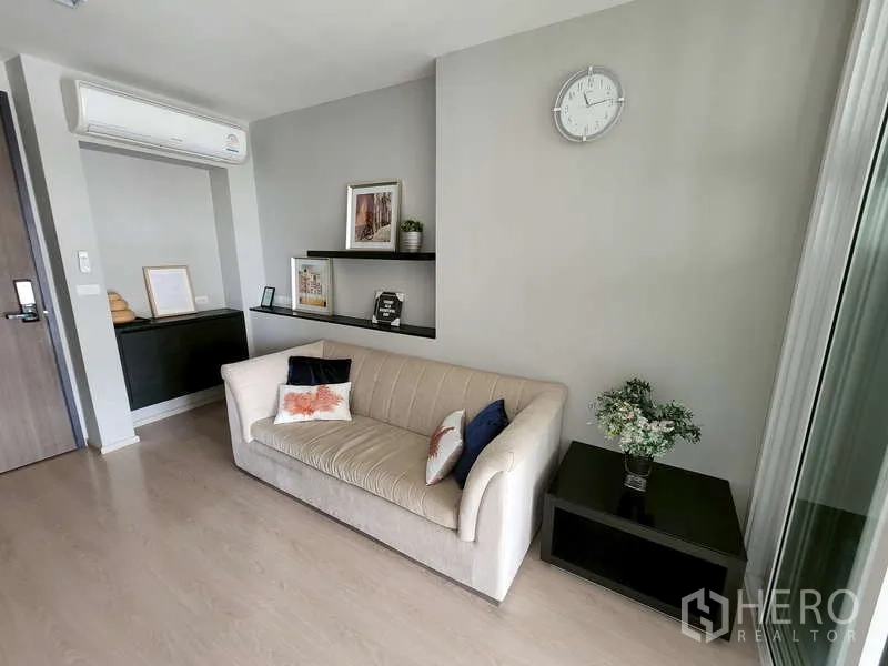 คอนโด ขาย - มุมนั่งเล่นอบอุ่นมีโซฟาสีเบจ ชั้นลอยตกแต่ง นาฬิกาแขวน และโต๊ะข้าง ใต้เครื่องปรับอากาศ