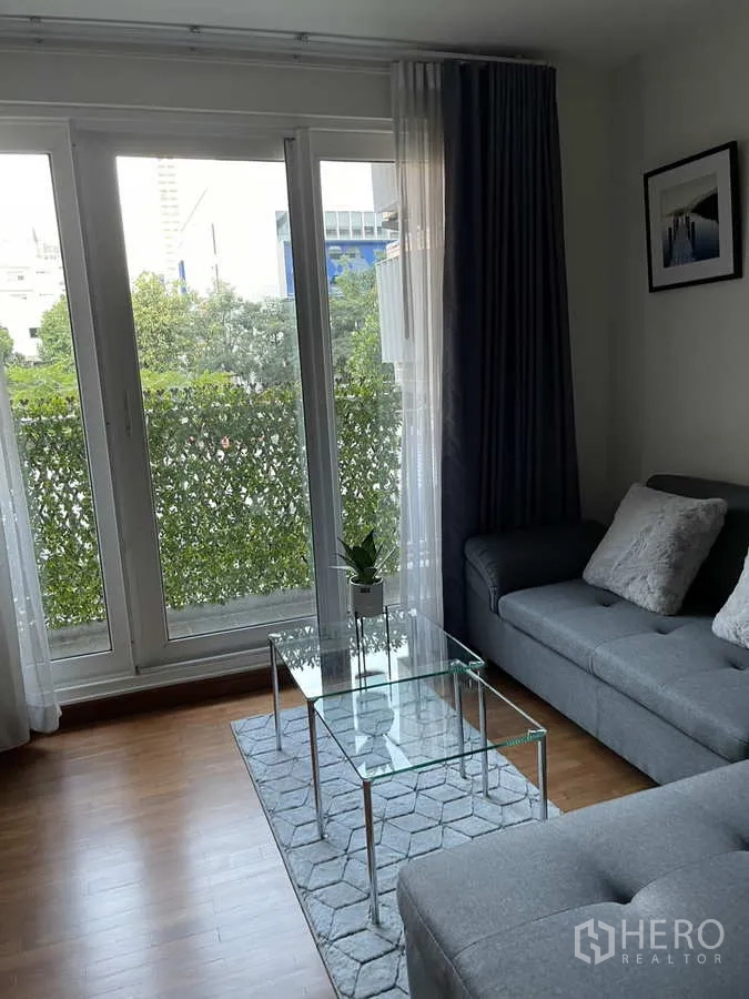 Condominium for rent in Watthana, Bangkok - Glass coffee table beside floor-to-ceiling balcony doors overlooking greenery.