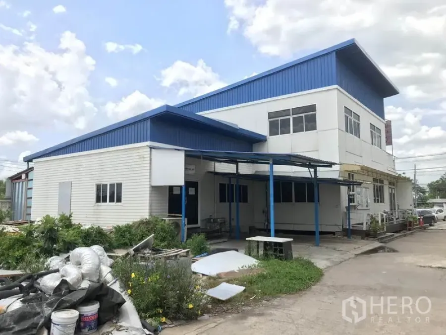 Warehouse for rent in Khlong Sam Wa, Bangkok - Two-storey warehouse with blue metal roof, attached office block and covered loading bay beside a wide driveway.