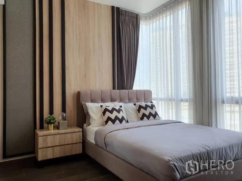 Condominium for rent or sale in Watthana, Bangkok - Master bedroom with tall corner windows, curtains and wood feature wall.