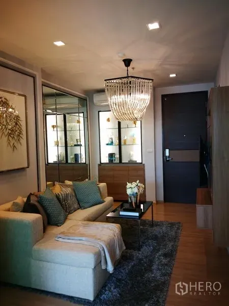 Condominium for rent in Sathon, Bangkok - Warm modern living room with L-shaped sofa, chandelier, mirrored display cabinet and wood flooring.