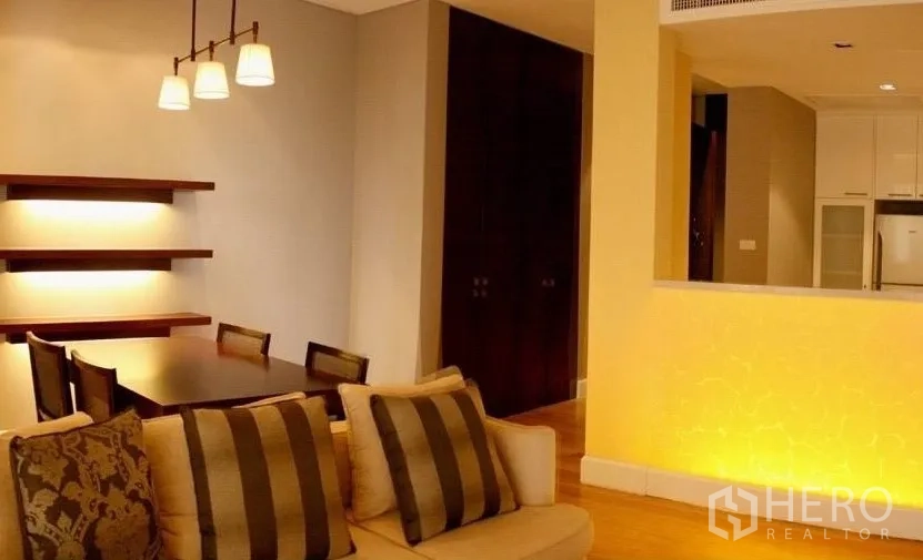 公寓 出租或出售在Watthana, Bangkok - 暖色照明的客餐厅，设沙发、餐桌、厨房传菜窗与壁挂层板。