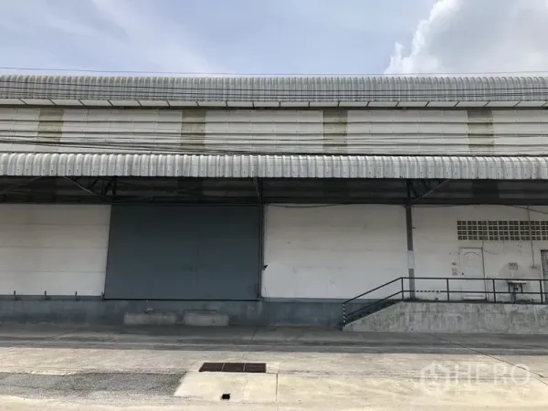 货栈 出租在Khlong Luang, Pathum Thani - 仓库外立面配大金属推拉门与坡道，屋檐雨棚覆盖。