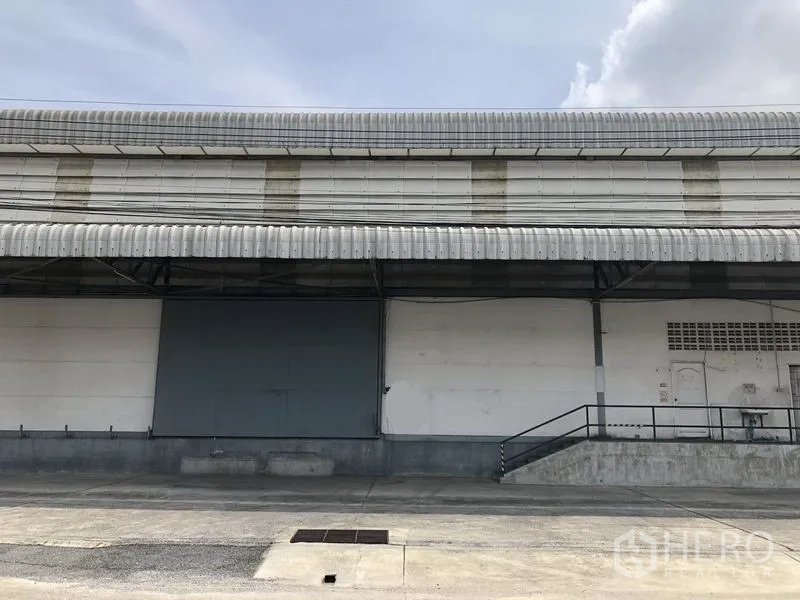 货栈 出租在Khlong Luang, Pathum Thani - 仓库外立面配大金属推拉门与坡道，屋檐雨棚覆盖。
