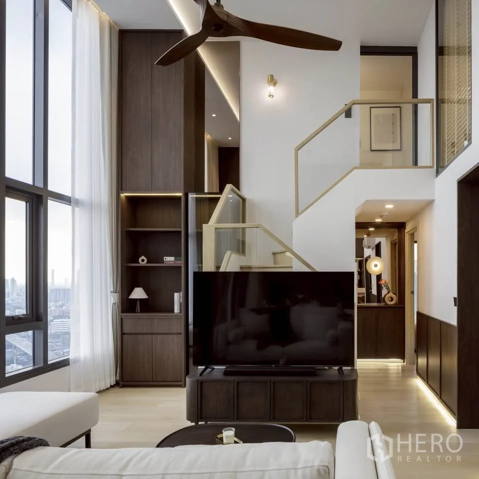 Condominium for rent in Khlong Toei, Bangkok - Double-height living room with staircase, tall windows, shelving and a ceiling fan.