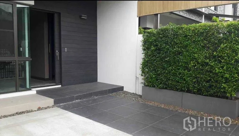 ทาวน์เฮาส์ ให้เช่า - ลานหน้าบ้านปูกระเบื้องสีเทา แนวรั้วต้นไม้ และประตูกระจกเข้าสู่ตัวบ้าน