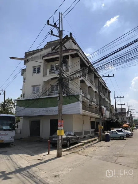 Shophouse for sale in Lam Luk Ka, Pathum Thani - Corner shophouse block on a main road with awnings, balconies, and power lines.