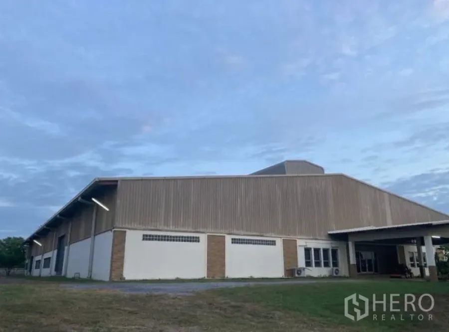 Factory for rent in Song Phi Nong, Suphan Buri - Large metal-clad factory exterior with covered loading bay and wide driveway on a grassy plot at dusk.
