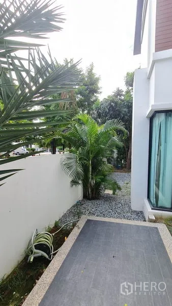 独立式住宅 出售在Samut Prakan - 房屋侧院种有棕榈，碎石小径与瓷砖露台。