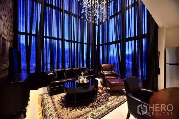 Condominium for rent or sale in Khlong Toei, Bangkok - Double-height living room with floor-to-ceiling curtains, crystal chandelier, leather sofa and classic rug at night.