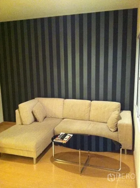 คอนโด ให้เช่าในWatthana, Bangkok - ห้องนั่งเล่น โซฟาเข้ามุมผ้าสีเบจ ผนังลายทางสีเข้ม และโต๊ะกลางกระจก พื้นไม้