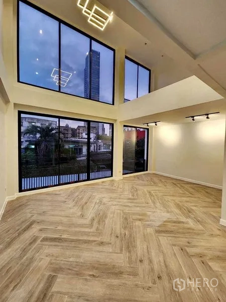 带办公室的家 出租或出售在Watthana, Bangkok - 双层挑高起居区，鱼骨拼木地板与整面落地窗眺望社区景观。
