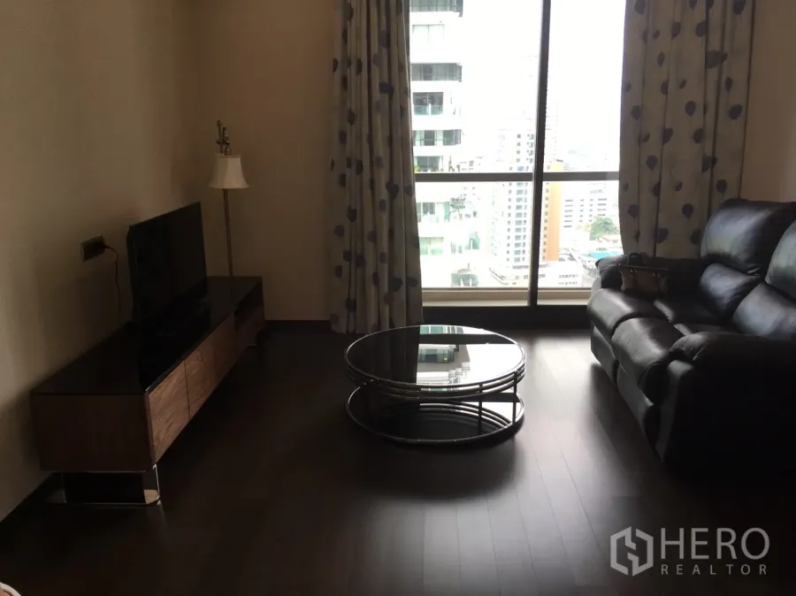 Condominium for rent in Watthana, Bangkok - Living room with black leather sofa, round glass coffee table, TV console, and floor-to-ceiling window showing city views.