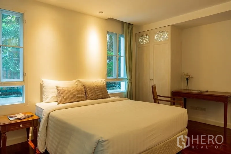 Apartment for rent in Bang Rak, Bangkok - Bright bedroom with double bed, side table and windows facing greenery, plus built‑in wardrobe and writing desk.