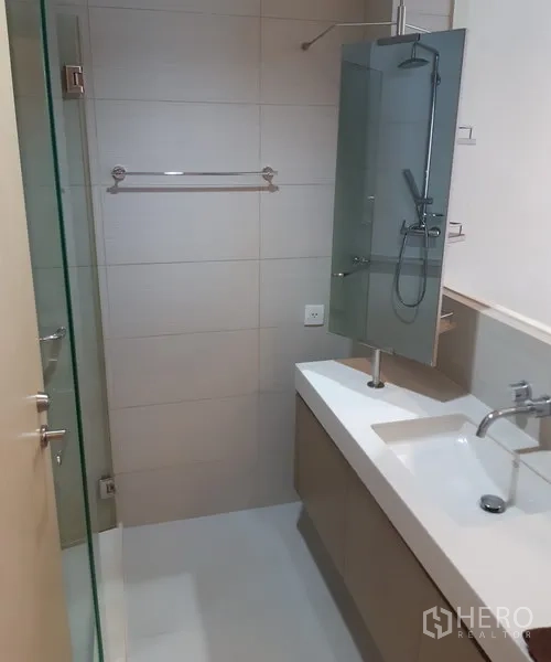 Condominium for rent in Khlong Toei, Bangkok - Contemporary bathroom with glass-enclosed shower, long vanity counter, and neutral wall tiles.