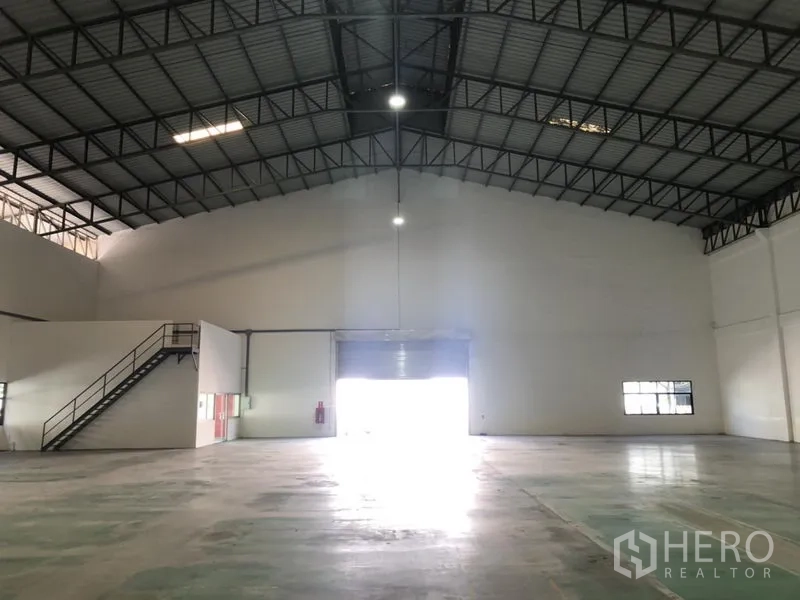 货栈 出租 - 宽敞仓库内景，高挑钢结构屋顶、卷闸门与夹层办公室。