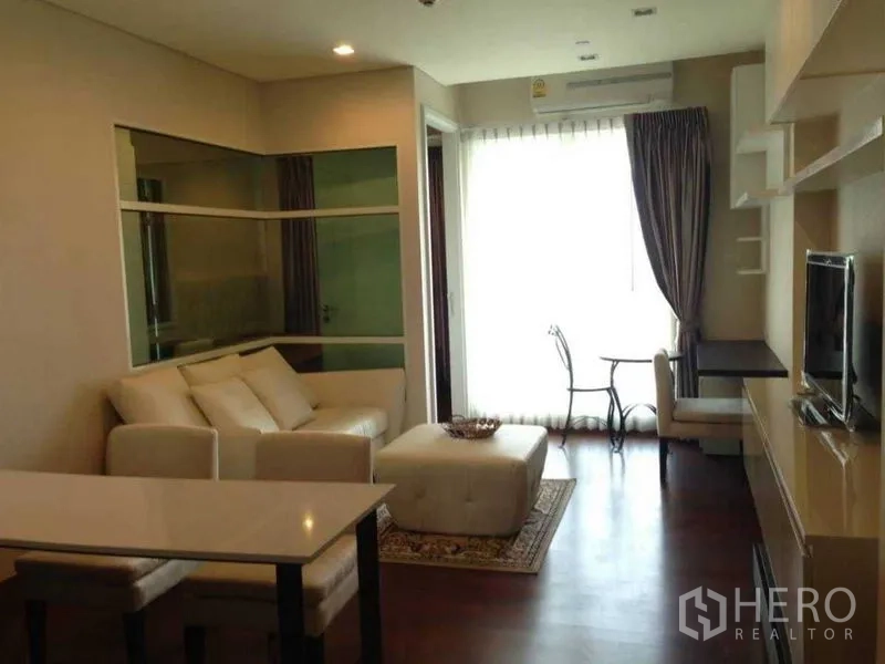 Condominium for rent or sale in Watthana, Bangkok - Bright living space with white sofa set, ottoman, balcony door with sheer curtains, wall-mounted TV and work desk.