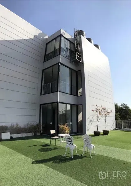 พื้นที่ขายปลีก ให้เช่าในBang Kapi, Bangkok - อาคารสมัยใหม่สีขาวหน้าต่างบานใหญ่ มองเห็นสนามหญ้าบนดาดฟ้าพร้อมที่นั่ง