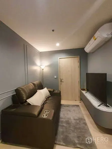 Condominium for rent - Compact lounge with leather sofa, floor lamp, wall-mounted TV console, and air conditioner.