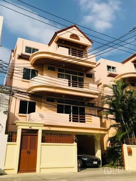 Detached House for rent in Watthana, Bangkok - Cream-colored multi-level detached house with balconies and a gated carport on a quiet lane.