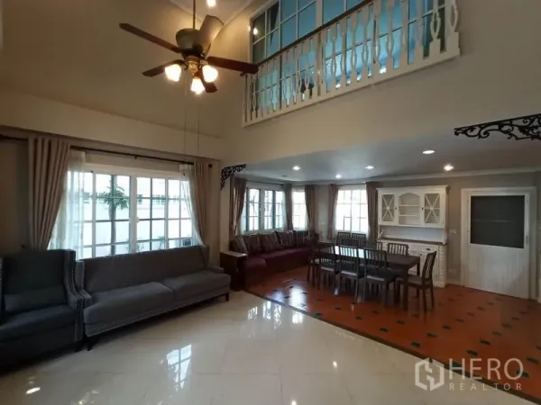 独立式住宅 出租在Mueang Samut Prakan, Samut Prakan - 挑高客厅，吊扇与大窗户，沙发区连通用餐区与楼中楼栏杆。