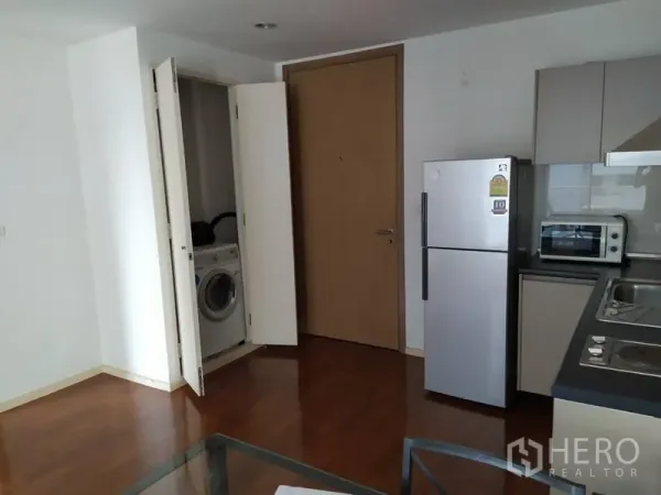 Condominium for sale in Khlong Toei, Bangkok - Open-plan kitchen with built-in cabinets, fridge, microwave and a closet laundry with washing machine on wood floors.