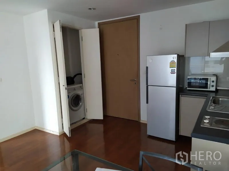 Condominium for sale in Khlong Toei, Bangkok - Open-plan kitchen with built-in cabinets, fridge, microwave and a closet laundry with washing machine on wood floors.