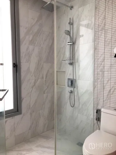Condominium for rent or sale in Watthana, Bangkok - Modern bathroom with glass-enclosed shower, rain showerhead and marble-look wall tiles.