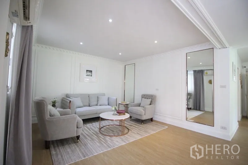 Detached House for rent in Chatuchak, Bangkok - Light living room with neutral sofas, armchair, round coffee table, large wall mirror, and recessed lighting.
