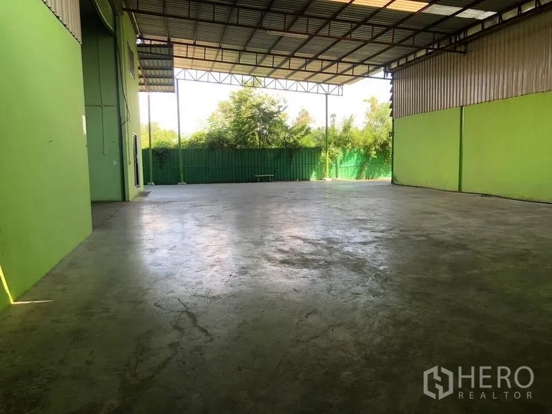 Factory for rent in Sam Khok, Pathum Thani - Covered open bay with concrete floor facing a green fence and trees.