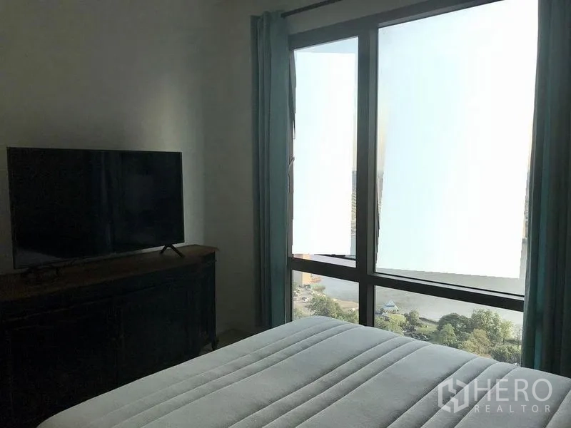 คอนโด ให้เช่าในBang Kho Laem, Bangkok - ห้องนอนมีหน้าต่างบานใหญ่ เห็นวิวเมืองและแม่น้ำ พร้อมเตียงและทีวีบนตู้สีเข้ม.