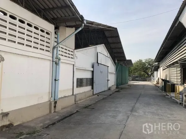 货栈 出租在Mueang Pathum Thani, Pathum Thani - 仓库建筑之间的外部通道，带大型滑门、金属屋顶和宽阔混凝土车道，适合卡车进出，位于Mueang Pathum Thani。