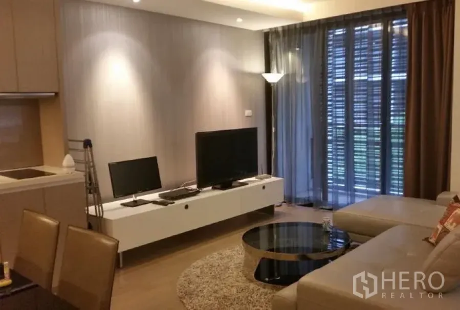 Condominium for rent or sale in Watthana, Bangkok - Modern living room with L‑shaped sofa, TV console, open‑plan kitchen and floor‑to‑ceiling windows leading to a balcony.