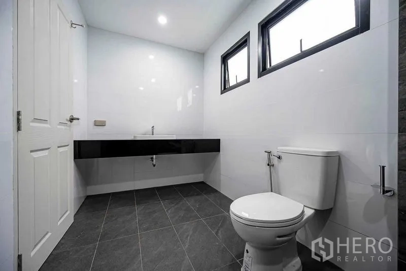 Home Office for rent in Suan Luang, Bangkok - Clean restroom with two high windows, a toilet, and a long black vanity counter.