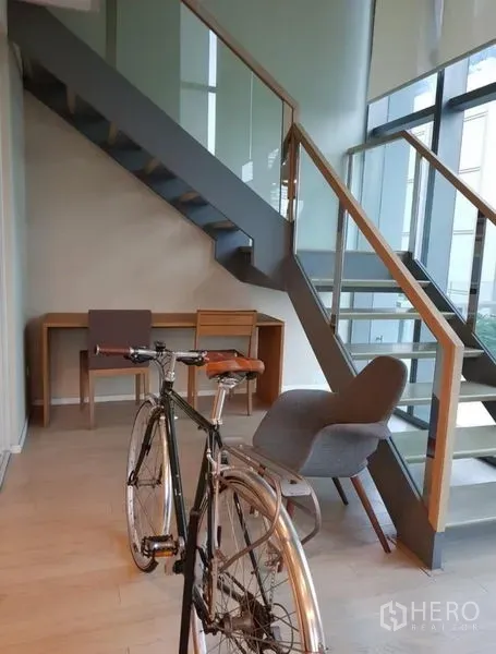 Condominium for rent or sale in Watthana, Bangkok - Work nook under the stairs with desk and lounge chair beside full-height windows and balcony access.