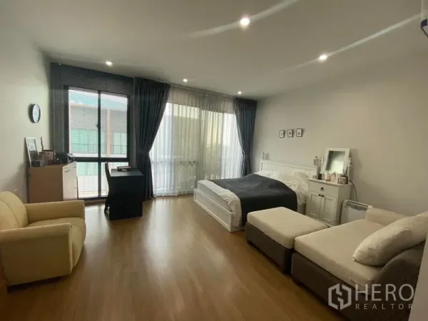 Townhouse for rent or sale in Saphan Sung, Bangkok - Spacious master bedroom with bed, sofa, wood floors and floor-to-ceiling curtains opening to a balcony.