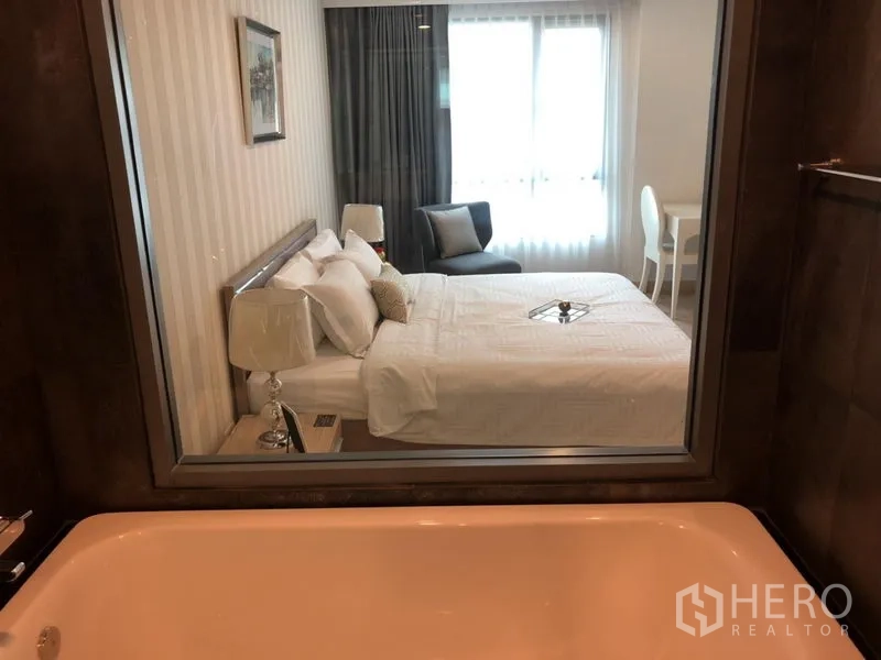 Condominium for rent in Watthana, Bangkok - Bright master bedroom viewed from the bathroom with a soaking tub, glass partition, bed, seating, and large window.