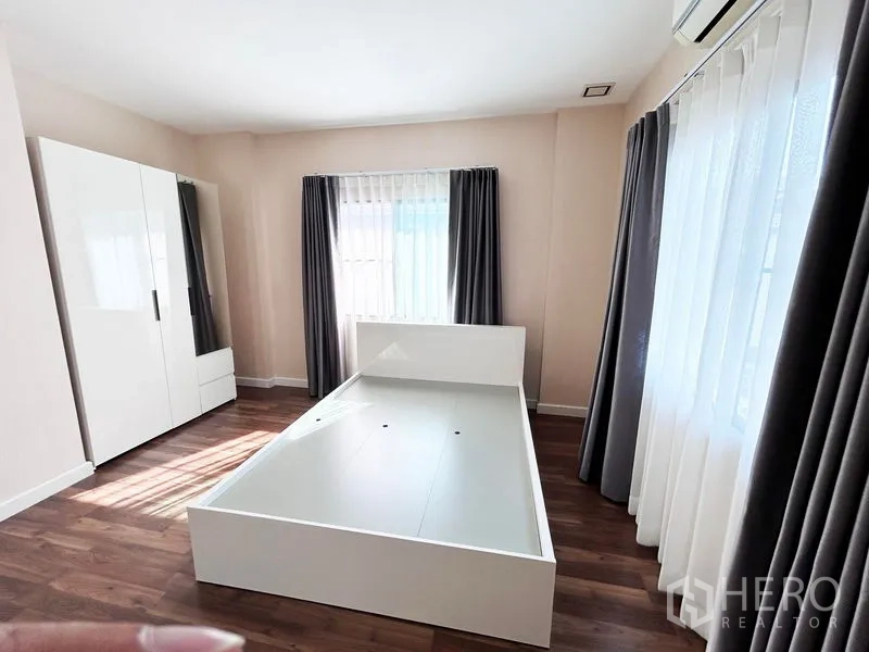 Detached House for rent or sale in Prawet, Bangkok - Bedroom with white bedframe, wardrobe, and full-height curtains.