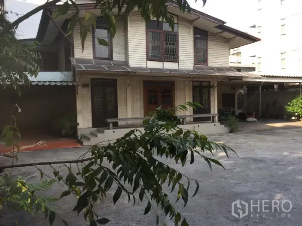 Detached House for rent or sale in Watthana, Bangkok - House exterior viewed past tree branches with expansive paved yard for many cars.